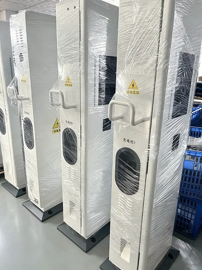 Batch of custom floor standing sheet metal DC EV charging station housings wrapped in protective film inside manufacturing workshop, reserved cutouts for charging gun and touch screen